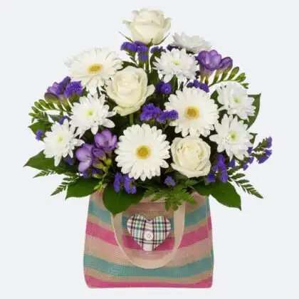 A vibrant arrangement of white roses, gerberas, and purple flowers in gift bag with a decorative heart.