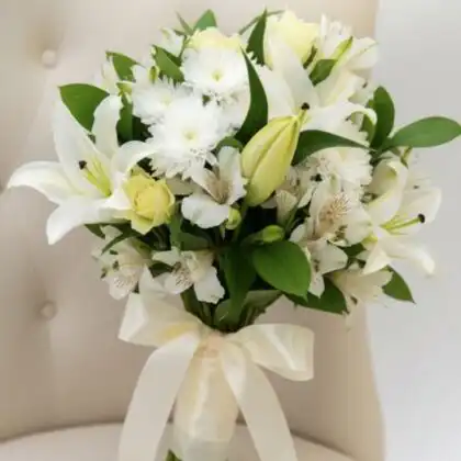 Hand-tied alstroemeria and lily bouquet with lush green leaves and a silk cream ribbon against a neutral background.